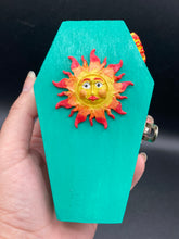 Load image into Gallery viewer, This coffin-shaped box features a bright turquoise exterior, with a bold yellow, orange and red sun adorning the center. Complemented with a shimmery rose-gold ribbon and flower, the inside is painted in a vibrant orange hue with turquoise crescent moon detailing and traditional Mexican-themed fabric. Coffin Dimensions: Length: 4 1/2 Inches; Width: 2 1/2 inches; Height: 1 5/8 Inches