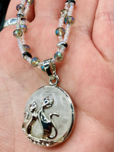 Load image into Gallery viewer, This stylish necklace features a shimmery sterling silver quartz pendant with cats and stars, making it the perfect accessory for any cat lover. With two-hole silver and black beads that stand out against the light aquamarine glass beads, this double strand necklace holds a round kitty pendant made with various hues of clear and iridescent glass beads. Add a touch of purrfection to your look with this statement piece. Measurements: Necklace is 15”adjustable up to 18"