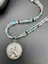 Load image into Gallery viewer, This stylish necklace features a shimmery sterling silver quartz pendant with cats and stars, making it the perfect accessory for any cat lover. With two-hole silver and black beads that stand out against the light aquamarine glass beads, this double strand necklace holds a round kitty pendant made with various hues of clear and iridescent glass beads. Add a touch of purrfection to your look with this statement piece. Measurements: Necklace is 15”adjustable up to 18"