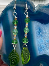 Load image into Gallery viewer, Long strand of glittering light green and gold glass beads, as well as a leaf-shaped glass charm at the bottom, combine to create a highly dynamic effect. These versatile earrings, perfect for a whimsical fairy princess, good witch, or nature-loving hippie look, draw on nature-inspired designs, providing the ideal finishing touch to any ensemble. Measurements: 2 1/4” in length