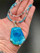 Load image into Gallery viewer, This Vibrant Sea Blue Agate Natural Stone Pendant Choker Style Beaded Necklace includes two strands of glass beads in shades of blues, silver and clear to lend shimmer and color to this large stone pendant. The shorter strand consists of very light blue small glass beads, while the longer strand is primarily a light baby blue. A set of dainty matching earrings completes the look. This beautiful gemstone necklace is the perfect accessory to bring the beach with you in the coming months.