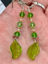 Load image into Gallery viewer, Long strand of glittering light green and gold glass beads, as well as a leaf-shaped glass charm at the bottom, combine to create a highly dynamic effect. These versatile earrings, perfect for a whimsical fairy princess, good witch, or nature-loving hippie look, draw on nature-inspired designs, providing the ideal finishing touch to any ensemble. Measurements: 2 1/4” in length