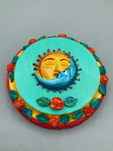 Load image into Gallery viewer, Handcrafted polymer clay wall art with a wood frame, featuring a sun and moon motif complemented with contrasting cool and warm colors. The background is a bright turquoise hue, with marigold flowers and leaves adorning the frame.Measurement 4" diameter