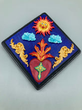 Load image into Gallery viewer, This handcrafted polymer clay wall art is displayed on a square wood frame, featuring sun and light blue cloud motifs on each side and a dark royal blue backdrop with a bright red sacred heart in the center. A turquoise and yellow cross provides contrast to the warm toned heart, which is further embellished with gold accents on each side of the heart. H 6 3/4" x W 6 3/4” x D 1"