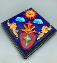 Load image into Gallery viewer, This handcrafted polymer clay wall art is displayed on a square wood frame, featuring sun and light blue cloud motifs on each side and a dark royal blue backdrop with a bright red sacred heart in the center. A turquoise and yellow cross provides contrast to the warm toned heart, which is further embellished with gold accents on each side of the heart. H 6 3/4" x W 6 3/4” x D 1"