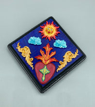 Load image into Gallery viewer, This handcrafted polymer clay wall art is displayed on a square wood frame, featuring sun and light blue cloud motifs on each side and a dark royal blue backdrop with a bright red sacred heart in the center. A turquoise and yellow cross provides contrast to the warm toned heart, which is further embellished with gold accents on each side of the heart. H 6 3/4" x W 6 3/4” x D 1"