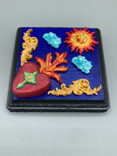 Load image into Gallery viewer, This handcrafted polymer clay wall art is displayed on a square wood frame, featuring sun and light blue cloud motifs on each side and a dark royal blue backdrop with a bright red sacred heart in the center. A turquoise and yellow cross provides contrast to the warm toned heart, which is further embellished with gold accents on each side of the heart. H 6 3/4" x W 6 3/4” x D 1"