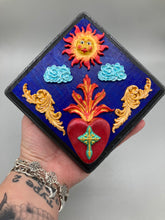 Load image into Gallery viewer, This handcrafted polymer clay wall art is displayed on a square wood frame, featuring sun and light blue cloud motifs on each side and a dark royal blue backdrop with a bright red sacred heart in the center. A turquoise and yellow cross provides contrast to the warm toned heart, which is further embellished with gold accents on each side of the heart. H 6 3/4" x W 6 3/4” x D 1"