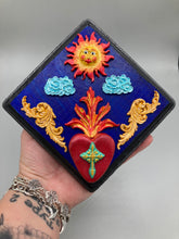 Load image into Gallery viewer, This handcrafted polymer clay wall art is displayed on a square wood frame, featuring sun and light blue cloud motifs on each side and a dark royal blue backdrop with a bright red sacred heart in the center. A turquoise and yellow cross provides contrast to the warm toned heart, which is further embellished with gold accents on each side of the heart. H 6 3/4" x W 6 3/4” x D 1"