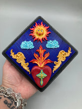 Load image into Gallery viewer, This handcrafted polymer clay wall art is displayed on a square wood frame, featuring sun and light blue cloud motifs on each side and a dark royal blue backdrop with a bright red sacred heart in the center. A turquoise and yellow cross provides contrast to the warm toned heart, which is further embellished with gold accents on each side of the heart. H 6 3/4" x W 6 3/4” x D 1"