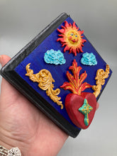 Load image into Gallery viewer, This handcrafted polymer clay wall art is displayed on a square wood frame, featuring sun and light blue cloud motifs on each side and a dark royal blue backdrop with a bright red sacred heart in the center. A turquoise and yellow cross provides contrast to the warm toned heart, which is further embellished with gold accents on each side of the heart. H 6 3/4" x W 6 3/4” x D 1"