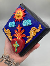 Load image into Gallery viewer, This handcrafted polymer clay wall art is displayed on a square wood frame, featuring sun and light blue cloud motifs on each side and a dark royal blue backdrop with a bright red sacred heart in the center. A turquoise and yellow cross provides contrast to the warm toned heart, which is further embellished with gold accents on each side of the heart. H 6 3/4" x W 6 3/4” x D 1"