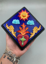 Load image into Gallery viewer, This handcrafted polymer clay wall art is displayed on a square wood frame, featuring sun and light blue cloud motifs on each side and a dark royal blue backdrop with a bright red sacred heart in the center. A turquoise and yellow cross provides contrast to the warm toned heart, which is further embellished with gold accents on each side of the heart. H 6 3/4" x W 6 3/4” x D 1"
