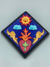Load image into Gallery viewer, This handcrafted polymer clay wall art is displayed on a square wood frame, featuring sun and light blue cloud motifs on each side and a dark royal blue backdrop with a bright red sacred heart in the center. A turquoise and yellow cross provides contrast to the warm toned heart, which is further embellished with gold accents on each side of the heart. H 6 3/4" x W 6 3/4” x D 1"