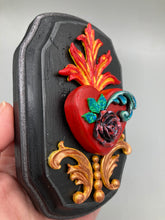 Load image into Gallery viewer, This Sacred Heart Wall Art Decor is the perfect way to enhance your home with an artistic flair. This beautiful artwork features a black wooden frame with a hand-crafted bright red heart. The heart is detailed with dark rose and vibrant green leaves, with a vibrant fire embellishment over the center. Gold decorative embellishments at the sides of the heart add a luxurious touch. H 5 3/8" x W 3 3/8” x D 1 1/2"