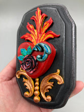 Load image into Gallery viewer, This Sacred Heart Wall Art Decor is the perfect way to enhance your home with an artistic flair. This beautiful artwork features a black wooden frame with a hand-crafted bright red heart. The heart is detailed with dark rose and vibrant green leaves, with a vibrant fire embellishment over the center. Gold decorative embellishments at the sides of the heart add a luxurious touch. H 5 3/8" x W 3 3/8” x D 1 1/2"