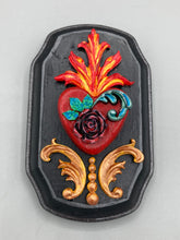 Load image into Gallery viewer, This Sacred Heart Wall Art Decor is the perfect way to enhance your home with an artistic flair. This beautiful artwork features a black wooden frame with a hand-crafted bright red heart. The heart is detailed with dark rose and vibrant green leaves, with a vibrant fire embellishment over the center. Gold decorative embellishments at the sides of the heart add a luxurious touch. H 5 3/8" x W 3 3/8” x D 1 1/2"