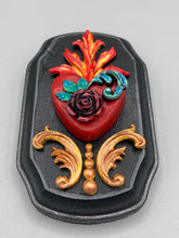 Load image into Gallery viewer, This Sacred Heart Wall Art Decor is the perfect way to enhance your home with an artistic flair. This beautiful artwork features a black wooden frame with a hand-crafted bright red heart. The heart is detailed with dark rose and vibrant green leaves, with a vibrant fire embellishment over the center. Gold decorative embellishments at the sides of the heart add a luxurious touch. H 5 3/8" x W 3 3/8” x D 1 1/2"