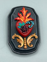 Load image into Gallery viewer, This Sacred Heart Wall Art Decor is the perfect way to enhance your home with an artistic flair. This beautiful artwork features a black wooden frame with a hand-crafted bright red heart. The heart is detailed with dark rose and vibrant green leaves, with a vibrant fire embellishment over the center. Gold decorative embellishments at the sides of the heart add a luxurious touch. H 5 3/8" x W 3 3/8” x D 1 1/2"