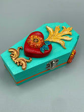 Load image into Gallery viewer, This coffin-shaped box features a bright turquoise exterior, with a hand-crafted bright red heart. The heart is detailed with a sunflower and vibrant green leaves, with a vibrant gold fire embellishment over the center. Gold decorative embellishments at the bottom of the heart add a luxurious touch. Complemented with a shimmery bright orange ribbon and flower around the coffin box, the inside is painted in a vibrant orange hue with black and silver cross and traditional Mexican-themed fabric.