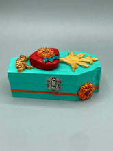 Load image into Gallery viewer, This coffin-shaped box features a bright turquoise exterior, with a hand-crafted bright red heart. The heart is detailed with a sunflower and vibrant green leaves, with a vibrant gold fire embellishment over the center. Gold decorative embellishments at the bottom of the heart add a luxurious touch. Complemented with a shimmery bright orange ribbon and flower around the coffin box, the inside is painted in a vibrant orange hue with black and silver cross and traditional Mexican-themed fabric.