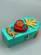 Load image into Gallery viewer, This coffin-shaped box features a bright turquoise exterior, with a hand-crafted bright red heart. The heart is detailed with a sunflower and vibrant green leaves, with a vibrant gold fire embellishment over the center. Gold decorative embellishments at the bottom of the heart add a luxurious touch. Complemented with a shimmery bright orange ribbon and flower around the coffin box, the inside is painted in a vibrant orange hue with black and silver cross and traditional Mexican-themed fabric.
