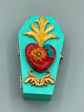 Load image into Gallery viewer, This coffin-shaped box features a bright turquoise exterior, with a hand-crafted bright red heart. The heart is detailed with a sunflower and vibrant green leaves, with a vibrant gold fire embellishment over the center. Gold decorative embellishments at the bottom of the heart add a luxurious touch. Complemented with a shimmery bright orange ribbon and flower around the coffin box, the inside is painted in a vibrant orange hue with black and silver cross and traditional Mexican-themed fabric.