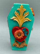 Load image into Gallery viewer, This coffin-shaped box features a bright turquoise exterior, with a hand-crafted bright red heart. The heart is detailed with a sunflower and vibrant green leaves, with a vibrant gold fire embellishment over the center. Gold decorative embellishments at the bottom of the heart add a luxurious touch. Complemented with a shimmery bright orange ribbon and flower around the coffin box, the inside is painted in a vibrant orange hue with black and silver cross and traditional Mexican-themed fabric.