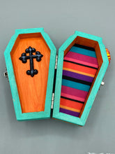 Load image into Gallery viewer, This coffin-shaped box features a bright turquoise exterior, with a hand-crafted bright red heart. The heart is detailed with a sunflower and vibrant green leaves, with a vibrant gold fire embellishment over the center. Gold decorative embellishments at the bottom of the heart add a luxurious touch. Complemented with a shimmery bright orange ribbon and flower around the coffin box, the inside is painted in a vibrant orange hue with black and silver cross and traditional Mexican-themed fabric.