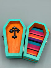 Load image into Gallery viewer, This coffin-shaped box features a bright turquoise exterior, with a hand-crafted bright red heart. The heart is detailed with a sunflower and vibrant green leaves, with a vibrant gold fire embellishment over the center. Gold decorative embellishments at the bottom of the heart add a luxurious touch. Complemented with a shimmery bright orange ribbon and flower around the coffin box, the inside is painted in a vibrant orange hue with black and silver cross and traditional Mexican-themed fabric.