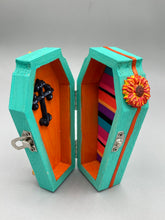 Load image into Gallery viewer, This coffin-shaped box features a bright turquoise exterior, with a hand-crafted bright red heart. The heart is detailed with a sunflower and vibrant green leaves, with a vibrant gold fire embellishment over the center. Gold decorative embellishments at the bottom of the heart add a luxurious touch. Complemented with a shimmery bright orange ribbon and flower around the coffin box, the inside is painted in a vibrant orange hue with black and silver cross and traditional Mexican-themed fabric.