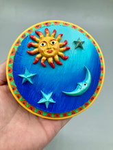 Load image into Gallery viewer, This handcrafted polymer clay wall art on a circle-shaped wood frame with a sun and crescent moon motifs on opposite side and stars. Its cool sky hues, including bright turquoise and dark royal blue, are further complemented by the bright orange and yellow frame adds an extra layer of sophistication to the warm-toned artwork. One of a kind art pieces come ready to hang with a sawtooth hangers in the back. Measurement 4" diameter.