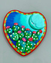Load image into Gallery viewer, This handcrafted polymer clay wall art features a heart-shaped wood frame complimented with a crescent moon motif. The complimentary color palette consists of cool sky hues, from a bright turquoise to a dark royal blue. The piece is further enhanced with hand sculpted cacti plants with blooming pink flowers. The vibrant red and orange frame complements the cool-toned artwork. Measurements: H 5 1/4" x W 5” x D 1 1/4"