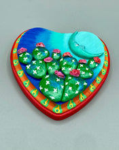 Load image into Gallery viewer, This handcrafted polymer clay wall art features a heart-shaped wood frame complimented with a crescent moon motif. The complimentary color palette consists of cool sky hues, from a bright turquoise to a dark royal blue. The piece is further enhanced with hand sculpted cacti plants with blooming pink flowers. The vibrant red and orange frame complements the cool-toned artwork. Measurements: H 5 1/4" x W 5” x D 1 1/4"