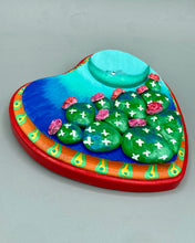 Load image into Gallery viewer, This handcrafted polymer clay wall art features a heart-shaped wood frame complimented with a crescent moon motif. The complimentary color palette consists of cool sky hues, from a bright turquoise to a dark royal blue. The piece is further enhanced with hand sculpted cacti plants with blooming pink flowers. The vibrant red and orange frame complements the cool-toned artwork. Measurements: H 5 1/4" x W 5” x D 1 1/4"