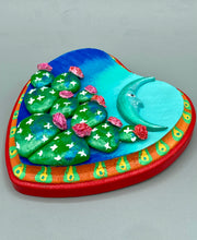 Load image into Gallery viewer, This handcrafted polymer clay wall art features a heart-shaped wood frame complimented with a crescent moon motif. The complimentary color palette consists of cool sky hues, from a bright turquoise to a dark royal blue. The piece is further enhanced with hand sculpted cacti plants with blooming pink flowers. The vibrant red and orange frame complements the cool-toned artwork. Measurements: H 5 1/4" x W 5” x D 1 1/4"