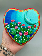 Load image into Gallery viewer, This handcrafted polymer clay wall art features a heart-shaped wood frame complimented with a crescent moon motif. The complimentary color palette consists of cool sky hues, from a bright turquoise to a dark royal blue. The piece is further enhanced with hand sculpted cacti plants with blooming pink flowers. The vibrant red and orange frame complements the cool-toned artwork. Measurements: H 5 1/4" x W 5” x D 1 1/4"