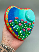 Load image into Gallery viewer, This handcrafted polymer clay wall art features a heart-shaped wood frame complimented with a crescent moon motif. The complimentary color palette consists of cool sky hues, from a bright turquoise to a dark royal blue. The piece is further enhanced with hand sculpted cacti plants with blooming pink flowers. The vibrant red and orange frame complements the cool-toned artwork. Measurements: H 5 1/4" x W 5” x D 1 1/4"