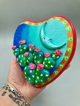 Load image into Gallery viewer, This handcrafted polymer clay wall art features a heart-shaped wood frame complimented with a crescent moon motif. The complimentary color palette consists of cool sky hues, from a bright turquoise to a dark royal blue. The piece is further enhanced with hand sculpted cacti plants with blooming pink flowers. The vibrant red and orange frame complements the cool-toned artwork. Measurements: H 5 1/4" x W 5” x D 1 1/4"