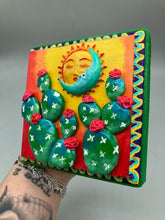 Load image into Gallery viewer, This handcrafted polymer clay wall art boasts a square-shaped wood frame with a sun and crescent moon motifs. Its warm sky hues, including yellow, orange and dark red, are further complemented by hand sculpted cacti plants with cool green-blue tones and dark pink flowers. The dark green frame adds an extra layer of sophistication to the warm-toned artwork. One of a kind art pieces come ready to hang with a sawtooth hangers in the back. H 5" x W 5” x D 1 1/2"H 5" x W 5” x D 1 1/2"