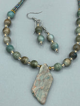 Load image into Gallery viewer, This exquisite necklace showcases earthy tones of mossy green ceramic beads, complemented by a gray toned howlite stone chip pendant. To add a sparkle, I interspersed gold-tone glass beads between each ceramic one. Two delicate strands feature small glass beads with hues of blue, gold, and purple. These are bridged by flower-shaped gold plated beads, which then unite with the round ceramic beads. This alluring earth-tone combination makes a stunning accessory. Necklace is 17”