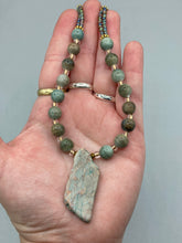 Load image into Gallery viewer, This exquisite necklace showcases earthy tones of mossy green ceramic beads, complemented by a gray toned howlite stone chip pendant. To add a sparkle, I interspersed gold-tone glass beads between each ceramic one. Two delicate strands feature small glass beads with hues of blue, gold, and purple. These are bridged by flower-shaped gold plated beads, which then unite with the round ceramic beads. This alluring earth-tone combination makes a stunning accessory. Necklace is 17”