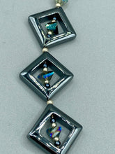 Load image into Gallery viewer, This two-strand necklace, crafted from aqua-blue and silver toned glass beads, is a stunning statement piece. The addition of faceted triangle-shaped glass beads tucked between hematite square beads adds an exquisite and unique flair. The necklace gracefully drapes around the neck, bringing an air of bold and elegant flair to any ensemble it complements. 15” in length, can be extended up to 17"
