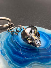 Load image into Gallery viewer, This beautiful edgy gun metal faceted shimmery multi-color glass skull bead keychain is full of hues of silver, purple, pink, blue, and gold. The split nickel key ring is 1 1/4" in diameter and the entire key chain is 4 1/2" in length (from key ring to skull bead)  Measurements: Nickel Free Key Ring is 1 1/4β in diameter, Entire Key Chain is 4 1/2" in Length, Metal Skull Bead 5/8" in Height