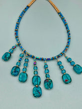 Load image into Gallery viewer, This necklace features vibrant teal quartzite and glass beads. The quartzite beads have a captivating swirl pattern, combining dark red and dark blue hues. It has an ethereal feel thanks to the various shades of blues, golds, and greens used to create a two-strand beaded design. For a dramatic finish, seven strands are added, completed with rectangular rainbow hematite beads for texture and depth. Both strands are held together with double-hole flat matte gold beads to keep a collar shape.
