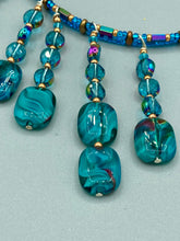 Load image into Gallery viewer, This necklace features vibrant teal quartzite and glass beads. The quartzite beads have a captivating swirl pattern, combining dark red and dark blue hues. It has an ethereal feel thanks to the various shades of blues, golds, and greens used to create a two-strand beaded design. For a dramatic finish, seven strands are added, completed with rectangular rainbow hematite beads for texture and depth. Both strands are held together with double-hole flat matte gold beads to keep a collar shape.
