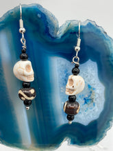 Load image into Gallery viewer, These mini mismatched earrings feature a howlite skull bead and two round wood carved beads. One bead features a circular and dot eye design, while the other displays a dot and a wave pattern, both in white. The wood beads have a dark brown hue, and black glass beads complete the look. Perfect for any goth, emo, or witchy look or to give your Dia De Los Muertos outfit an original feel. Measurements: 1 1/2 ” in length