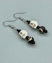 Load image into Gallery viewer, These mini mismatched earrings feature a howlite skull bead and two round wood carved beads. One bead features a circular and dot eye design, while the other displays a dot and a wave pattern, both in white. The wood beads have a dark brown hue, and black glass beads complete the look. Perfect for any goth, emo, or witchy look or to give your Dia De Los Muertos outfit an original feel. Measurements: 1 1/2 ” in length