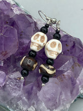 Load image into Gallery viewer, These mini mismatched earrings feature a howlite skull bead and two round wood carved beads. One bead features a circular and dot eye design, while the other displays a dot and a wave pattern, both in white. The wood beads have a dark brown hue, and black glass beads complete the look. Perfect for any goth, emo, or witchy look or to give your Dia De Los Muertos outfit an original feel. Measurements: 1 1/2 ” in length