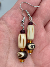Load image into Gallery viewer, This set of earrings features a cylinder bone bead and round bone bead with eye design and a gold-tone glass bead. Neutral colors allow for versatile styling; the resulting look is sure to be unique.  Measurements: 1 1/2 ” in length